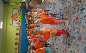 dzieci w pomarańczowych strojach z emblematami marchewki na opaskach na głowie, maszerują po dywanie. Jeen z chłopców niesie pomarńczowy koszyk