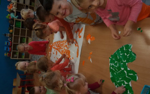 dzieci zgromadzone dookoła dużego stołu wyklejają kontur marchewki na brystolu