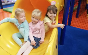 trzy usmiechnięte dziewczynki siedzą u wylotu wielkiej, żółtej zjeżdżalni
