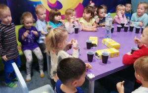 dzieci na małych kzesełkach przy fioletowym stole piją sok z kubeczków i zajadają ciastka