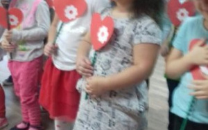 na scenie dzieci w czerwonych czapkach, z czerwonym sercem ozdobionym białym kwiatuszkiem w dłoniach