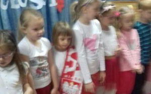 Grupka dzieci w biało-czerwonych strojach stoi na scenie z rękami opuszczonymi wzdłuż tułowia