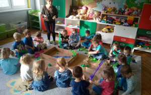 dzieci na dywanie siedzą dookoła szarych arkuszy papieru na których leży niskie, kartonowe pudło. rozwijają szerokie pasy bibuły