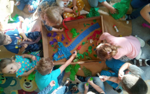 dzieci pochylone nad kartonowym pudłem, przygotowują makietę puszczy - wklejeją do kartonu gniecioną, formowaną dłońmi bibułę - rośliny, woda, kwiaty, drzewa