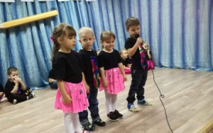 dwie dziewczynki w różowych spódniczkach oraz dwóch chłopców w tęczowych krawatach, stoją na scenie. jeden z chłopców trzyma mikrofon
