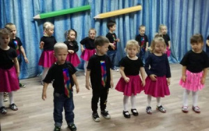 dzieci w czarnych tshirtach, chłopcy w tęczowych krawatach, dziewczynki w różowych spódniczkach, stoją w rozsypce na scenie i śpiewają piosenkę