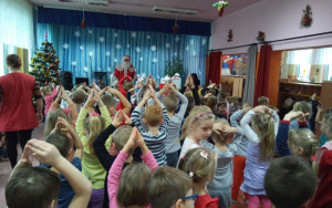 Mikołaj przed sceną, śpiewa i tańczy razem z dziećmi, ramiona wyciągnięte nad głowami dzieci tworzą daszek - złączone palce