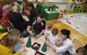 rodzice wraz z dziećmi z dużym zaangażowaniem ozdabiają zielone choinki kolorowymi ozdobami, w tle stół ze słodkościami