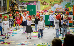 3 latki w odświetnych strojach i czerwonych czapeczkach krasnali, śpiewa na dywanie, kilkoro dzieci wyciąga ramiona do góry