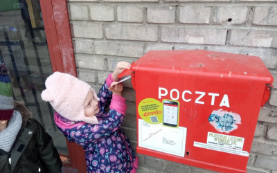dziewczynka w kolorowej kurtce i różowej czapce z uszkami wrzuca kartkę do czerwonej skrzynki pocztowej