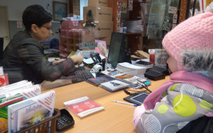 dziewczynka w różowej czapeczce obserwuje panią w okienku pocztowym, która przybija stempel na świątecznej kartce