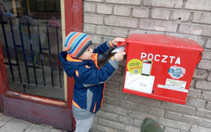 chłopiec w granatowej kurtce i czapce w paski wrzuca kartkę do czerwonej skrzynki pocztowej