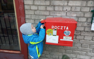 chłopiec w niebieskiej kurtce i szarej czapce wrzuca kartkę do czerwonej skrzynki pocztowej