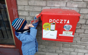 chłopiec w niebieskiej kurtce i czapce w paski wrzuca kartkę do czerwonej skrzynki pocztowej