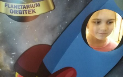 ziewczynka pozuje do zdjęcia w okrągłym oienku rakiety, z lewej strony napis Planetarium Orbitek