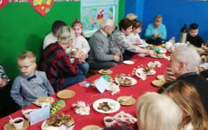 Babcie i dziadkowie z wnukami przy stole przykrytym czerwonym obrusem i zastawionym słodkościami