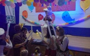 na scenie za girlandą z balonów tańczy bałwanek Olaf, przed sceną tańczą dorośli z białymi pałeczkami w rękach i dzieci