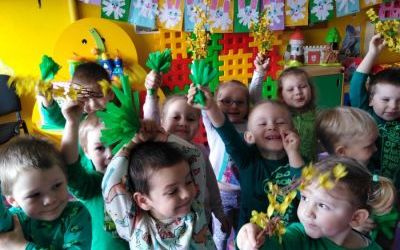 przedszkolaki machają radośnie zieloną bibułkową trawką oraz żółtymi forsycjami