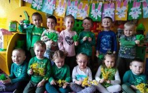 dzieci pozują do zdjęcia ze sztucznymikwiatami forsycji oraz bibułkowymi, zielonymi trawkami w dłoniach